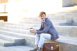 © PhotoStar - Young man in a stylish suit sitting on outdoor steps, holding a coffee cup, with a leather briefcase beside him, enjoying a moment of relaxation in a modern urban setting