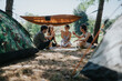 © qunica.com - A group of friends camping in a wooded campsite share snacks and conversation under a tarp shelter, enjoying a relaxed outdoor moment together in warm daylight.