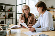 © AnnaStills - Two middle aged Caucasian women discussing documents at desk, collaborating on business project with laptops and charts, focused on reviewing paperwork in modern office setting