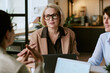 © AnnaStills - Caucasian middle aged woman wearing glasses engaging in business meeting with two colleagues in modern office setting, discussing project while sitting at table with laptops visible