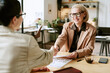 © AnnaStills - Middle aged Caucasian woman smiling while shaking hands with another middle aged woman across desk in modern office setting, discussing documents and business collaboration