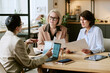 © AnnaStills - Three Caucasian middle aged women sitting at table discussing business documents and analyzing charts with laptop and tablet in modern office setting during meeting