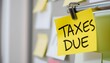 © Husen - A bright yellow sticky note with the words 'TAXES DUE' written in black marker, attached to a metal bar with a clip.
