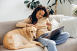 © Maria Vitkovska - Authentic African American woman sitting on comfortable sofa using laptop petting golden retriever
