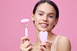 © SHOTPRIME STUDIO - Young woman showcasing facial skincare tools on a pastel pink background, promoting healthy skin routines with a joyful expression