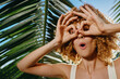 © SHOTPRIME STUDIO - Young woman with curly hair making binocular gesture with hands near tropical palm leaves in bright sunlight, playful mood, summer outdoor fun and natural background.