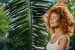 © SHOTPRIME STUDIO - smiling woman with curly hair enjoying tropical sunlight beside large green palm leaf in summer outdoor setting, natural beauty and happiness captured.