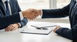 © Farhan - Two businessmen in navy suits shake hands over a white desk after signing a contract, symbolizing a finalized legal agreement and successful negotiation.