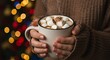 © Joverth - Close-up of hands holding a mug of hot cocoa with marshmallows, Christmas lights blurred in the background, cozy festive mood