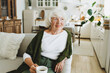 © shurkin_son - Smiling stylish senior lady with grey hair enjoying her freshly brewed coffee sitting in relaxed pose on sofa in her tidy cozy living-room looking aside. Happy retirement and longevity concept