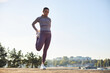 © DragonImages - Young adult Caucasian woman stretching leg outdoors on waterfront, preparing for workout, standing on one foot with hand holding ankle, trees and buildings visible in background