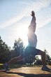 © DragonImages - Young adult Caucasian man performing outdoor yoga stretching exercise on wooden platform with arms raised overhead, sunlight shining through trees in background, wearing athletic shoes