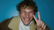 © SerPak - A cheerful young man with curly hair and glasses smiles broadly while making a peace sign. The background is a vibrant blue, creating a joyful atmosphere.