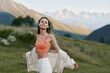 © SHOTPRIME STUDIO - A cheerful woman wearing an orange sports top stands outdoors in a scenic valley with mountains in the background, smiling as she embraces the sunshine and fresh air.