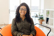 © Prostock-studio - A cheerful young woman with curly hair and glasses sits comfortably in a vibrant orange chair. Sunlight streams through large windows, illuminating the stylish room filled with greenery.