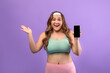 © Prostock-studio - A happy young european plus size woman in sportswear stands against a purple background. She shows a blank phone screen with excitement, promoting a potential amazing offer for a sports app.