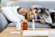 © sofiko14 - A young child rests on a sofa, appearing unwell, with various medications placed on a table in the foreground, suggesting illness.