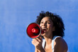 © VISTA by Westend61 - Woman with afro hairstyle shouting through red megaphone by blue wall