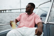 © TrueFrame Collective - Black young adult man wearing sunglasses relaxing on boat holding cold drink with straw, sitting by water with bridge in background, enjoying leisure time outdoors