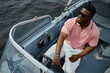 © TrueFrame Collective - Black young adult man steering motorboat on water, wearing sunglasses and looking away from camera, sitting in driver seat with hand on wheel, enjoying boating activity