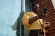 © TrueFrame Collective - Black young adult man standing outdoors holding smartphone in one hand and disposable coffee cup in other hand, smiling while looking at mobile device near glass building