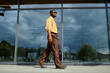 © TrueFrame Collective - Young adult Black man walking confidently outdoors in front of modern glass building, wearing sunglasses, looking ahead with relaxed posture, urban environment reflected in windows