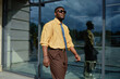 © TrueFrame Collective - Young adult Black man walking outdoors near glass building, wearing sunglasses and smiling, confident expression, modern urban setting reflected in background window