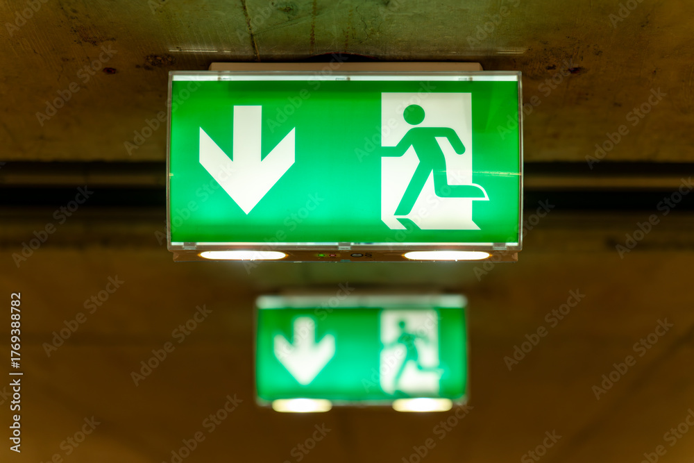 Two green emergency exit signs in a row, the background one blurry ...
