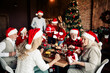 © deagreez - Family celebration at Christmas dinner with Santa hats around a festive table near a decorated tree and gifts
