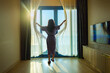 © shine - Morning of Pretty young woman opening curtains in new apartment. Morning light through the window in her room. Clear blue sky with a few clouds. Empty large white room with laminate flooring.