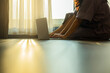 © shine - Woman freelancer working on laptop while sitting on floor. Light filters through sheer curtains, casting intricate shadows on a wooden floor.