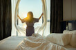 © shine - Confident middle aged woman opens the curtains with white flying curtains, looking at the view from window Beautiful sunrise above sea or ocean. Woman enjoying her holiday in a luxury hotel. Back view