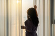© shine - Thoughtful middle aged woman opening curtain for beautiful sunshine, thinking about future in cozy apartment in moment of quiet introspection while drinking morning coffee by the window in bedroom.