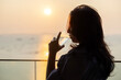 © shine - Confident asian middle aged woman drinking water from glass of water for future wellness. Sunrise sky clouds over sea in the morning with Orange sunlight. Concept of healthy diet. Copy space for ad.