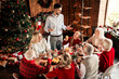 © deagreez - Family gathers for Christmas dinner by a decorated tree in a cozy home setting with lights and smiles