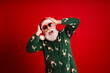© deagreez - Playful Santa in green Christmas pajama with reindeer pattern wearing sunglass against a bold red background for festive holiday vibes