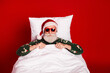 © deagreez - Santa relaxes in a cozy red bedroom wearing a festive pajama sweater and lounging in bed for a playful Christmas photo
