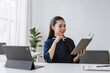 © amnaj - Asian businesswoman working, studying, and smiling, holding a notebook and stylus in a modern office with a tablet