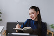 © amnaj - Young Asian woman writing in notebook or journal, sitting on sofa at home, studying or working remotely