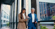 © serg - Confident business professionals in formal suits walking and talking outside a modern office building female executive using a digital tablet, male colleague holding a laptop
