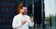 © serg - Attractive businessman wearing white shirt is on a video cal,l meeting conference influencer, entrepreneur business city while walking. He is holding his phone in one hand and use wireless headphones