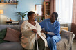 © DragonImages - Senior Black woman sitting on sofa holding cane, talking with young adult Black female nurse in medical scrubs, providing support and companionship in home setting