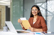 © Liubomir - Young indian woman smiling with excitement as she opens a letter at her desk in a modern office, celebrating a job offer or important career news while working on a laptop