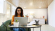 © Prostock-studio - A woman with curly hair and glasses sits in a cozy chair at a small table. She looks surprised while working on her laptop in a bright and stylish workspace.