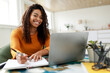 © Prostock-studio - A woman with curly hair smiles as she types on her laptop and writes notes in a notebook. Sunlight fills the cozy home office, creating a warm atmosphere for productivity.