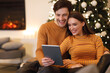 © Prostock-studio - A young couple shares a warm moment while looking at a tablet. They are seated comfortably near a Christmas tree adorned with decorations in a cozy living room setting.