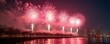 © Viktor - Pink fireworks explode over a city waterfront at night. Illuminated display reflects in water. Beautiful colorful show during festival or holiday event. Buildings silhouettes in background.