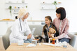 © sofiko14 - A female doctor consults with a young girl and her mother on a sofa, likely during a home visit. The child appears unwell, wrapped in a blanket and holding a teddy bear.