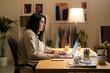 © Studio Marmellata - Positive young Arab woman in casual sweater sitting at table with laptop and working online on project from home during evening time