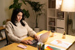 © Studio Marmellata - Positive young Arab woman in casual sweater sitting at table with laptop and working online on project from home during evening time